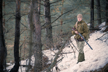 Female hunter in camouflage clothes ready to hunt, holding gun and walking in forest.