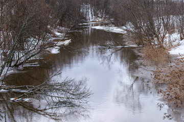 Naklejka na ściany i meble A dull gray landscape in early spring begins to melt snow and ice, a reflection in the river.