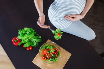 Pregnant woman in kitchen is eating vegetable salad.