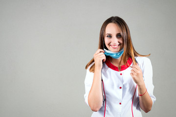 Red-haired girl or woman doctor or nurse with stethoscope on neck, on a light background in doctor's dressing gown looks into camera smiling and puts on antiviral gauze mask