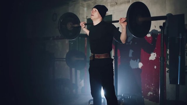 Muscular man doing crossfit training in a dark shadowy gym lifting weights holding a barbell at waist level in a health and fitness concept, movement round close shot.