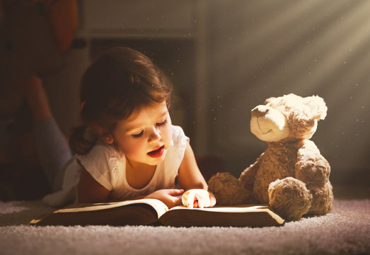 Little child girl is reading a book in evening in dark with a toy bear.