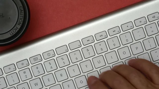 Elderly Man Is Typing On A Keyboard. On The Table Is A Cup Of Coffee. Red Background.