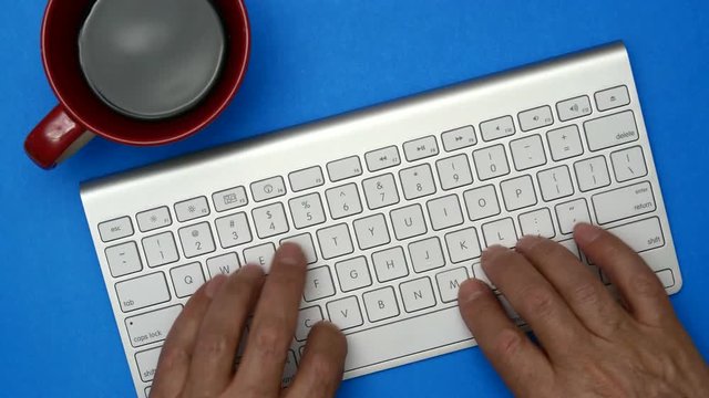 Elderly Man Is Typing On A Keyboard. On The Table Is A Cup Of Coffee. Blue Background.