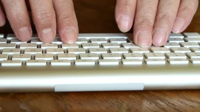 Elderly Man Is Typing On A Keyboard While Sitting At A Wooden Table.