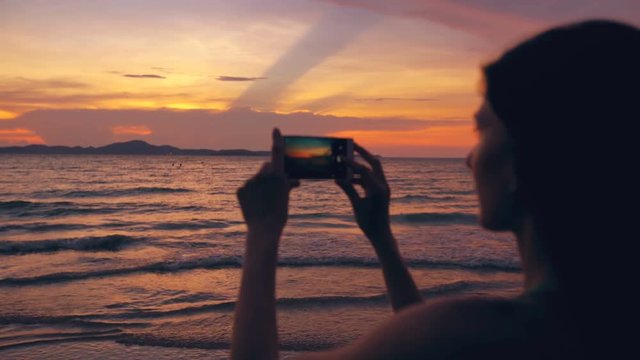 Silhouette of young tourist woman photographs ocean view with smartphone during sunset at beach