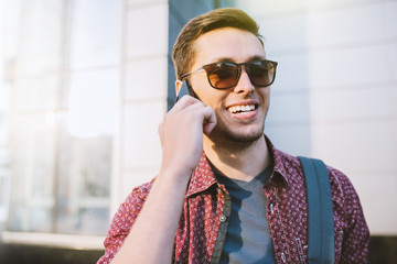 Cheerful attractive caucasian man in stylish sunglasses in the city. Portrait of young business man using mobile phone on building outdoors. Traveler man talking on smart phone on the street.