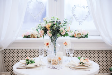 Interior design of white evening room with beautiful chandelier under served table with flowers on it. Big windows at the background. Vertical