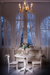 Interior design of white evening room with beautiful chandelier under served table with flowers on it. Big windows at the background. Vertical
