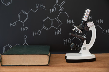 microscope and book on wooden table with science formula on black background