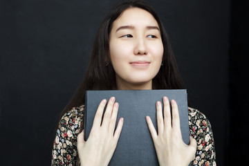 An asian girl in a dress is holding a black book in her hands. She looks dreamily away. Love of...