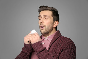 Young man sneezing in tissue on gray background