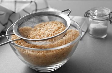 Sieve with raw brown rice in bowl on table