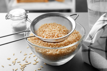 Sieve with raw brown rice in bowl on table