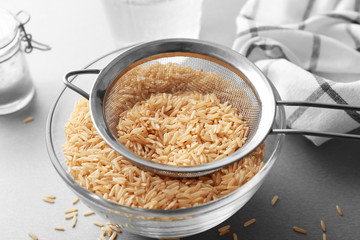 Sieve with raw brown rice in bowl on table