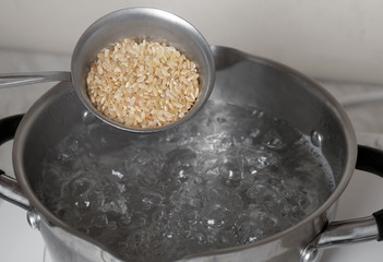 Raw brown rice in measuring cup over saucepan with boiling water