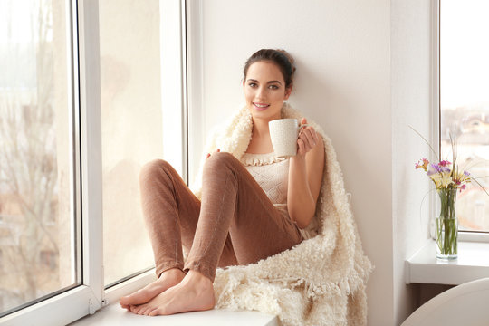Beautiful young woman drinking coffee while sitting on window sill at home