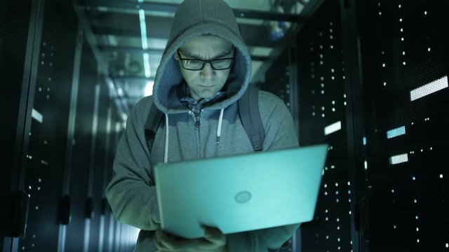 Hacker in a Hoodie Standing in the Middle of Data Center full of Rack Servers and Hacking it with His Laptop.  Shot on RED EPIC-W 8K Helium Cinema Camera.