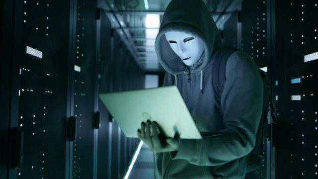Close-up Shot of a Masked Hacker in a Hoodie Standing in the Middle of Data Center full of Rack Servers and Hacking it with His Notebook.  Shot on RED EPIC-W 8K Helium Cinema Camera.