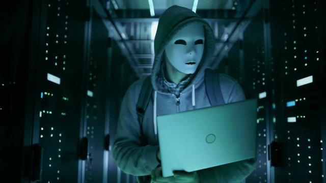 Close-up Shot of a Masked Hacker in Holding Laptop and Hacking Data Server.  Shot on RED EPIC-W 8K Helium Cinema Camera.