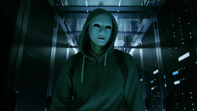 Masked Hacker in a Hoodie Walks Through Corporate Data Center with Rows of Working Rack Servers.  Shot on RED EPIC-W 8K Helium Cinema Camera.