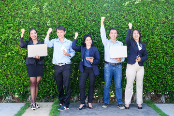 Group of business people arm up and holding lab-top computer, tablet, smart phone standing in front of green tree leaves background, Business triumph, teamwork concept