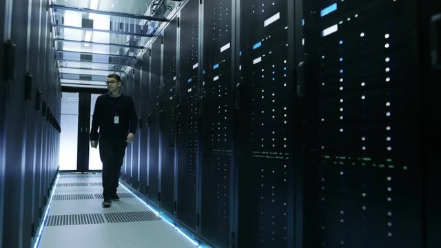 Frontal View of IT Engineer Holding Notebook and Walking Through Data Center Full of Working Rack Servers.  Shot on RED EPIC-W 8K Helium Cinema Camera.