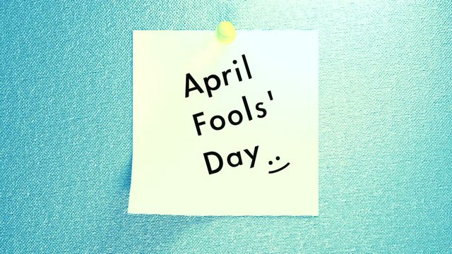 April Fools' Day note. Cartoon style happy holidays greetings animation. White sticky note with pin on fabric texture wall. Wriggling and jiggling text effects. Cute smiley face emoticon.