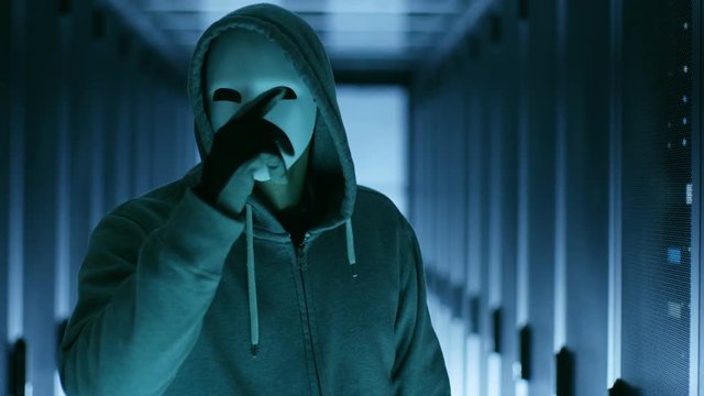 Masked Hacker Makes Threatening Gesture to the Camera. He is Standing in a Corridor full of Servers. Shot on RED EPIC-W 8K Helium Cinema Camera.