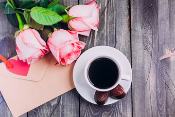 Holiday card. Pink roses with paper greeting card envelope and coffee cup with chocolate candy for Mothers day/Valentine's day on wooden table background. Copy space. Top view. Flat design.