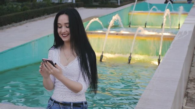 Girl laughs using smartphone. Beautiful young woman with her mobile phone on illuminated fountain background.