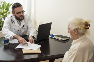 Lekarz geriatra przyjmuje pacjenta w gabinecie. Stara kobieta u lekarza geriatry. Lekarz tłumaczy pacjentce zapis w dokumentach. - obrazy, fototapety, plakaty