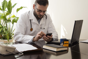 Lekarz w gabinecie lekarskim siedzi przy biurku z telefonem komórkowym w dłoni.Lekarz wybiera numer na telefonie. - obrazy, fototapety, plakaty