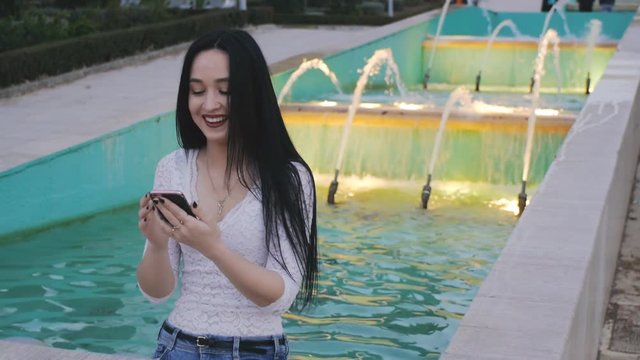 Girl laughs and flips hair using smartphone. Beautiful young woman with her mobile phone on illuminated fountain background.