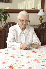 Stara kobieta ze smutną miną liczy pieniądze siedząc przy stole.Staruszka liczy pieniądze siedząc przy stole. - obrazy, fototapety, plakaty