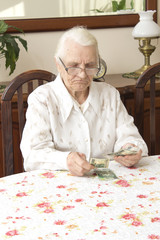 Emerytura. Stara kobieta liczy pieniądze. Stara kobieta ze smutną miną liczy pieniądze siedząc przy stole. - obrazy, fototapety, plakaty