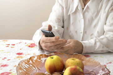Dłonie starej kobiety trzymającej telefon komórkowy. Babcia siedzi przy stole w salonie i trzyma w dłoniach telefon.