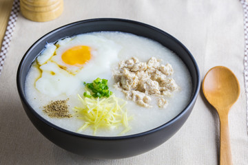 Close up to rice porridge is topping with ginger, spring onion, pepper, chicken, and onsen egg, Breakfast set on table, spoon, pepper grinder. breakfast in Asia call 