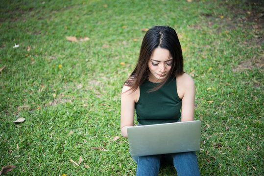 Young beautiful woman sitting on green grass and using laptop in the park with  nature background.