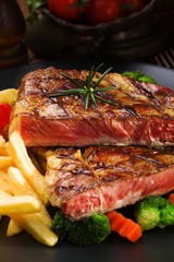 Grilled beef steak served with French fries and vegetables on a black plate.