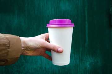 Mock up paper cup of coffee for branding .Cup of coffee with violet cap in woman's hand against green background. Good morning concept. Empty space for inscription.