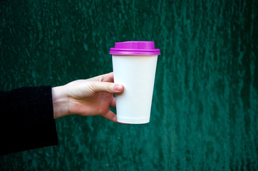 Mock up paper cup of coffee for branding .Cup of coffee with violet cap in woman's hand against green background. Good morning concept. Empty space for inscription.