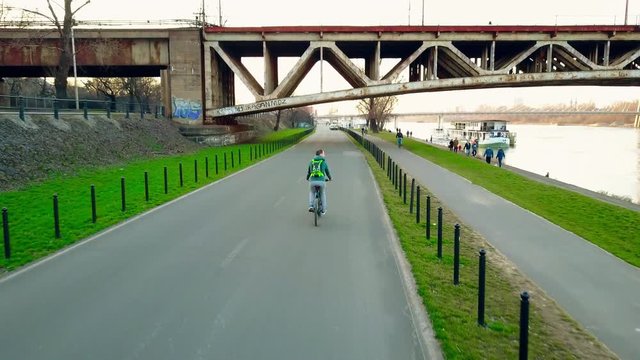 Aerial tracking shot of young man with backpack riding his bike along evening river embankment. 4K clip
