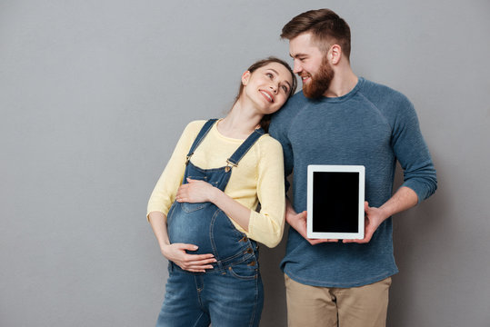 Cheerful couple expecting child and showing blank tablet computer screen