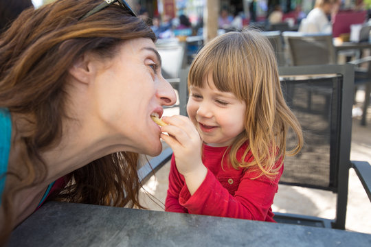 blonde three years old child face, with red shirt, feeding mother with cheese puff, laughing, sitting in terrace exterior bar cafe with grey table in park 
