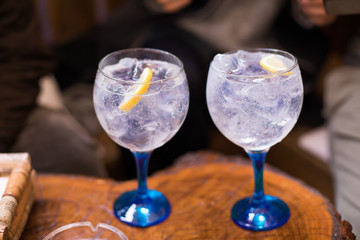 two transparent glass wide cups, with blue crystal, full of gin tonic cocktail drink, with lemon slice and ice cubes, on wooden table
