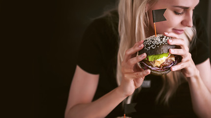 Blond girl in a black T-shirt with a big black burger in her hands