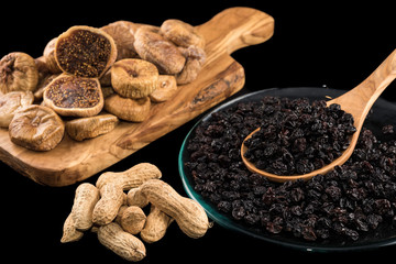 Dried figs on wooden chopping board, peanuts and black raisins in plate on black background