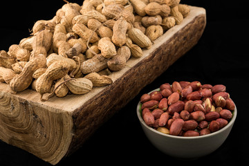 Peanuts in nutshell on a piece of wood and peeled peanuts in white bowl on black background