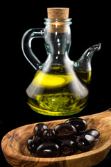 Black olives in wooden spoon and bottle of virgin olive oil on black background